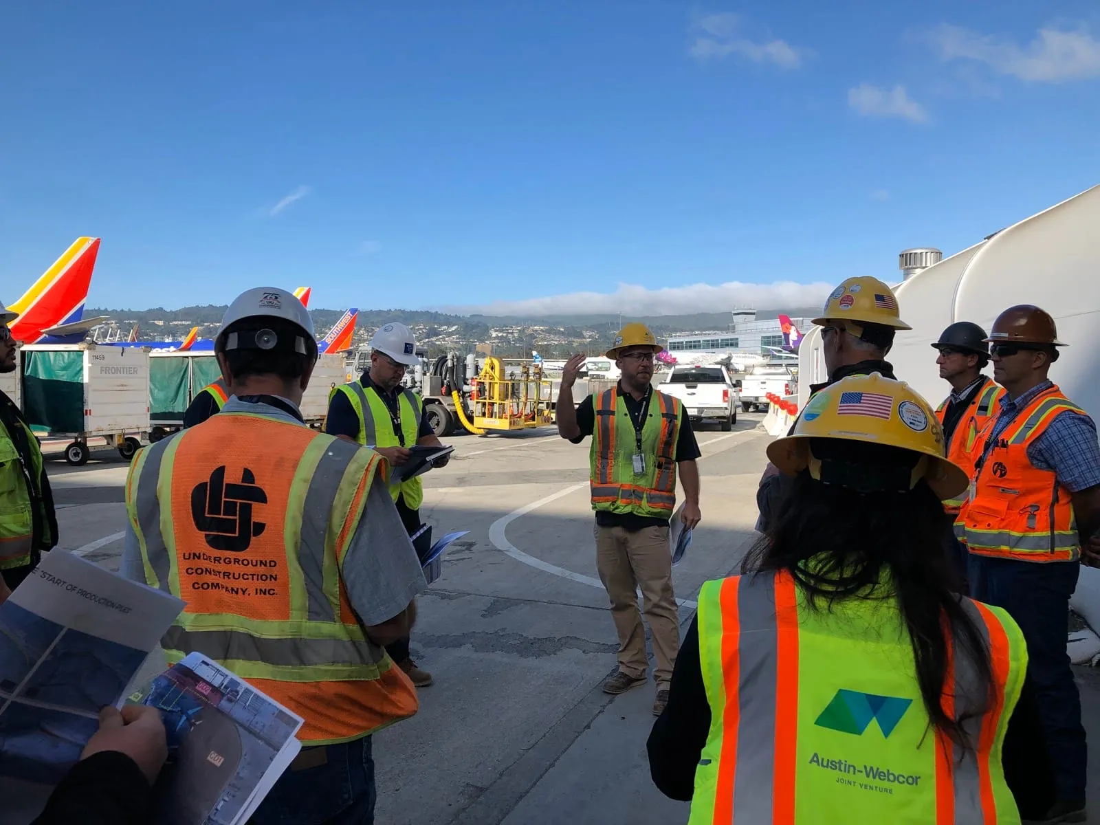A huddle of builders in vests on the SFO apron, jets parked behind them