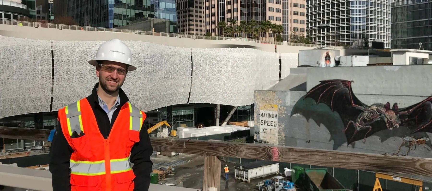 Chris in a hard hat and vest, the transit center’s white lattice skin behind him